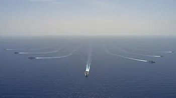 Seven naval ships sailing in formation in open water with the horizon behind them,